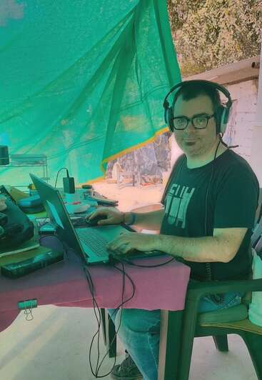 Um homem de óculos e fones de ouvido trabalha em um laptop ao ar livre, debaixo de uma lona verde, cercado por equipamentos eletrônicos sobre uma mesa coberta por um pano rosa. Ele está sorrindo.