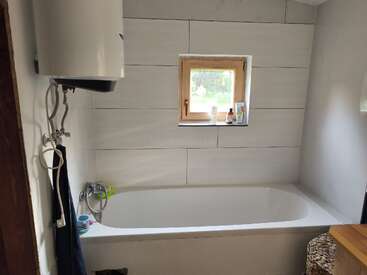 The image depicts a bathroom with a white bathtub, tiled walls, and a window, featuring a water heater, towel rack, and various toiletries on the windowsill.