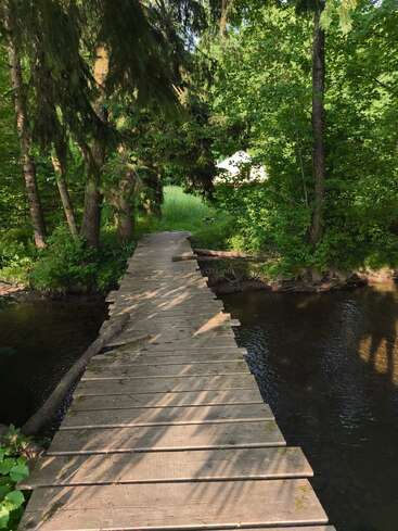 The image depicts a weathered wooden bridge spanning a river, surrounded by lush greenery and trees, with a serene and natural atmosphere.