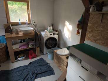 The image depicts a compact bathroom with a sink, toilet, and washing machine, featuring white walls and floors, and a window with a view of the outdoors.