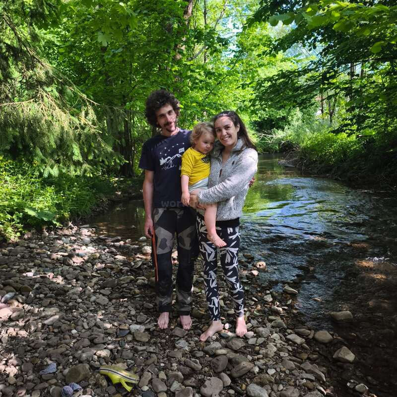 The image depicts a family of three standing on rocks near a serene river, surrounded by lush greenery and trees, with a peaceful atmosphere.
