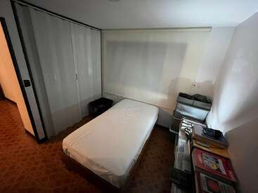 This image shows a small, tidy bedroom with a single bed, closet, window shade, stack of books, plastic storage drawers, and patterned tile flooring.