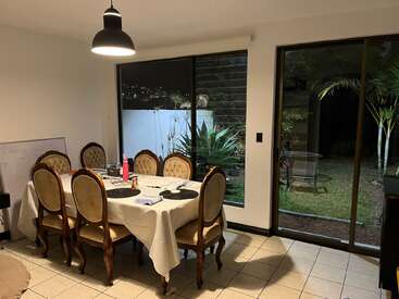A cozy dining room with a table set for six, vintage chairs, white tablecloth, modern hanging lamp, large windows reveal a garden and outdoor plants.