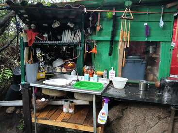 This image shows an outdoor kitchen with a makeshift sink, dish racks, utensils, cleaning supplies, hanging brushes, and shelves filled with cups and plates. Rustic setup.