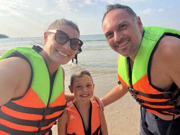 Das Bild zeigt eine dreiköpfige Familie mit neongrünen und orangefarbenen Schwimmwesten an einem Strand, die für ein Foto mit dem Meer im Hintergrund posiert.
