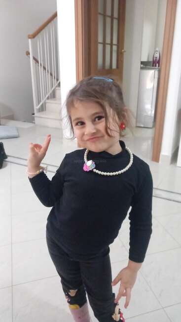 Ein junges Mädchen steht drinnen und lächelt total frech. Sie hat ein schwarzes Outfit an, dazu eine Perlenkette und bunte Ohrringe, und posiert selbstbewusst. Hinter ihr sieht man eine Treppe und eine Tür.