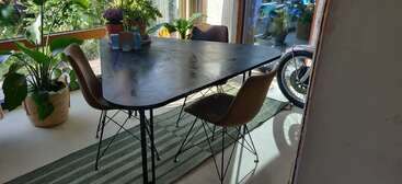 Une salle à manger moderne avec une table triangulaire noire, quatre chaises marron, diverses plantes en pot, un tapis rayé et une moto visible près de la fenêtre.