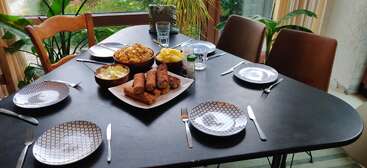 Une table à manger noire est dressée pour quatre personnes, avec des assiettes à motifs, des couverts, des boissons, des fritures et des chips, le tout entouré de plantes d'intérieur près de grandes fenêtres.