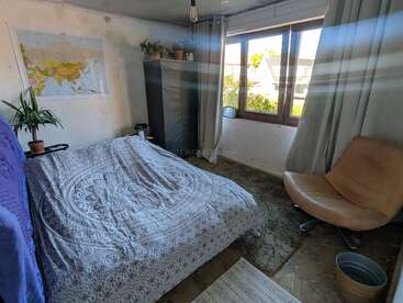 Une chambre confortable avec un couvre-lit à motifs, une grande fenêtre, une chaise marron, une armoire, des plantes, une carte du monde accrochée au mur et la lumière qui inonde la pièce.