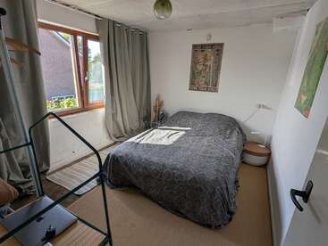 Une chambre confortable et baignée de soleil, avec des rideaux gris, un lit double recouvert d'une couverture sombre, des décorations murales, une fenêtre ouverte, une petite table de chevet ronde et une décoration minimaliste.