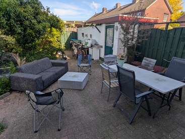 Une terrasse confortable dans le jardin comprend un canapé gris, des chaises pliantes, une grande table entourée de différentes chaises, des plantes vertes, un petit abri de jardin et des vélos en arrière-plan.
