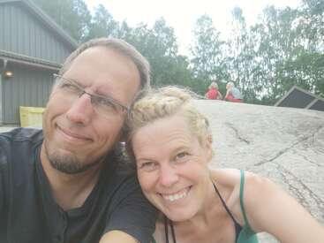 Das Bild zeigt ein Selfie von einem Mann und einer Frau, die zusammen im Freien posieren, mit einem Gebäude und Bäumen im Hintergrund. Sie lächeln beide.