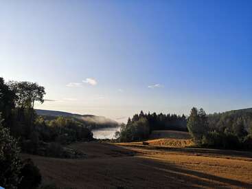 Das Bild zeigt eine ruhige Landschaft mit einem Feld, Bäumen und einem See, mit einem klaren blauen Himmel und einem sanften Nebel, der von der Wasseroberfläche aufsteigt.
