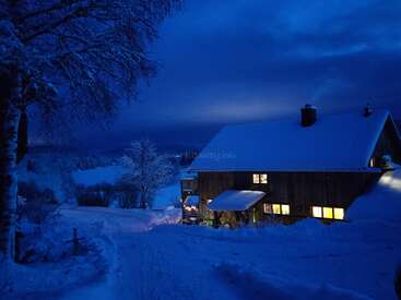 Ein gemütliches Holzhaus mit schneebedecktem Dach und erleuchteten Fenstern, eingebettet in eine ruhige Schneelandschaft unter einem dunkelblauen Nachthimmel.