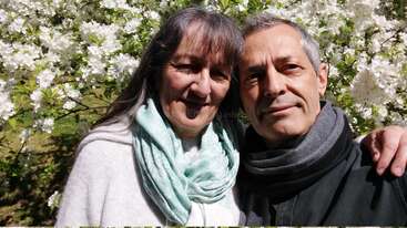 Un couple âgé se tient serré l'un contre l'autre, le sourire aux lèvres. Ils se trouvent en plein air, entourés de fleurs blanches en pleine floraison ; tous deux portent un foulard et profitent d'une journée ensoleillée.