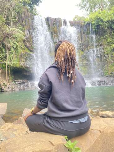 Eine Person mit Dreadlocks sitzt auf einem Felsen und blickt auf einen wunderschönen Wasserfall, der von üppigem Grün umgeben ist und eine friedliche, ruhige Szene neben dem fließenden Wasser schafft.