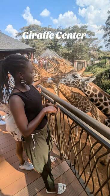 Eine Frau steht auf einer Plattform im Giraffe Center und füttert große Giraffen. Andere Besucher sind zu sehen. Blauer Himmel, Bäume und verspielte Giraffen umgeben sie.