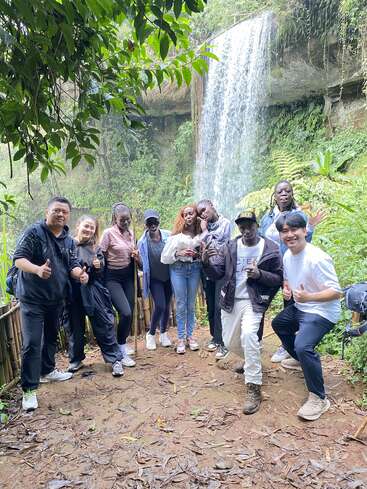 Eine bunt gemischte Gruppe von Freunden posiert fröhlich vor einem wunderschönen Wasserfall inmitten von üppigem Grün und fängt ein lustiges Outdoor-Abenteuer in der Natur ein.