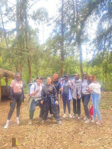 Eine Gruppe von acht jungen Leuten posiert zusammen in einem sonnenbeschienenen Wald, lächelnd und entspannt, lässig gekleidet, ein lustiges Outdoor-Abenteuer zwischen hohen grünen Bäumen genießend.