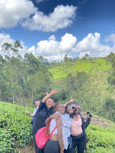 Eine Gruppe von Freunden posiert fröhlich zwischen üppigen grünen Teesträuchern auf einem Hügel, mit strahlend blauem Himmel, flauschigen Wolken und dichtem Wald im Hintergrund.
