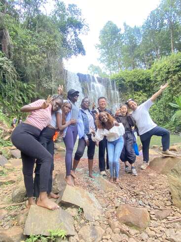Eine fröhliche Gruppe von Freunden posiert barfuß auf Felsen vor einem wunderschönen Wasserfall im Wald, lächelnd, lachend und mit Friedenszeichen in einer üppigen Umgebung.