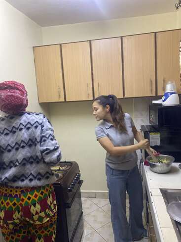 Zwei Frauen sind in einer kleinen Küche beschäftigt. Eine Frau rührt Zutaten in einer Schüssel um, während die andere neben dem Herd steht und Essen zubereitet.