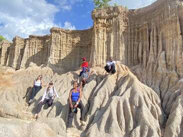 Cinco personas posan sobre impresionantes formaciones rocosas erosionadas con majestuosos y escarpados acantilados a sus espaldas. El cielo es brillante y azul, creando una escena dramática al aire libre.