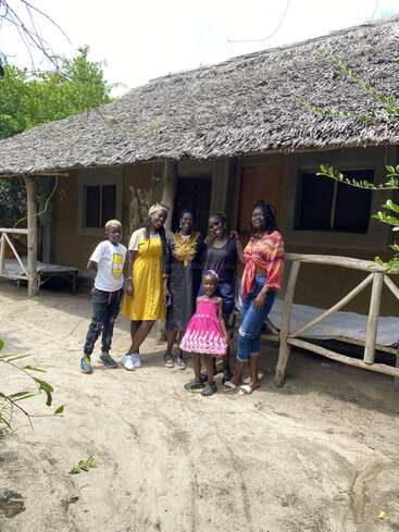 Six people, including a young child, stand smiling in front of a rustic, thatched-roof house surrounded by greenery. The group appears happy and relaxed outdoors.