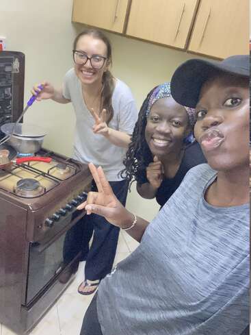 Tres mujeres sonríen en una cocina. Una está cocinando en un fogón, mientras las otras dos posan para un selfie, mostrando signos de paz. Ambiente amistoso.