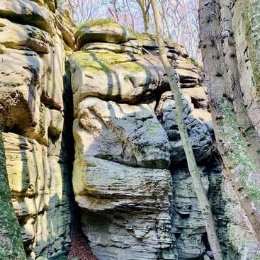 L'image représente une formation rocheuse accidentée, recouverte de mousse et de lichen, avec une fissure verticale proéminente, entourée d'arbres et sur fond de ciel bleu vif.