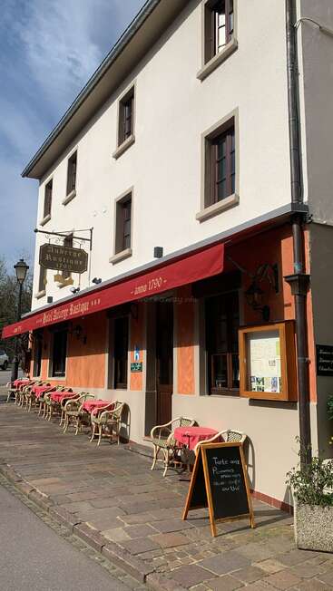 L'image montre un bâtiment de deux étages avec un auvent rouge et un espace de restauration en plein air, avec un panneau indiquant "Auberge Rustique" et un tableau noir avec une écriture en français.