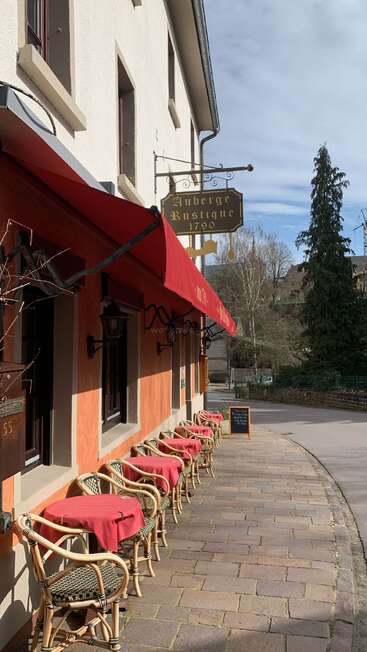 L'image représente un charmant espace extérieur à l'extérieur d'un bâtiment avec un auvent rouge, avec des tables et des chaises, et un panneau indiquant "Auberge Rustique 1790" en lettres dorées.