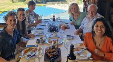 L'image représente un groupe de personnes réunies autour d'une table, probablement en train de prendre un repas ensemble. La scène se déroule dans une salle à manger ou à l'extérieur.