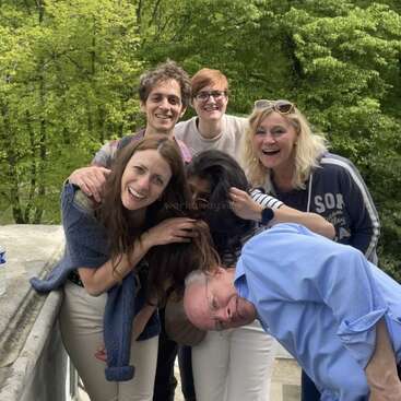 L'image représente un groupe de six personnes posant pour une photo, avec un homme penché et les autres l'embrassant, sur fond d'arbres.