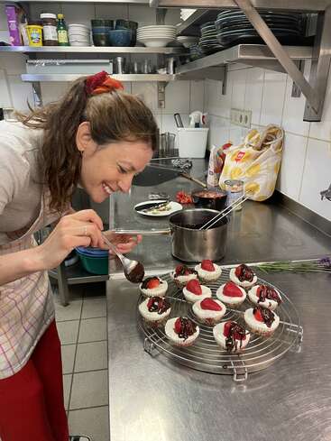 Une femme dans une cuisine décore de petits gâteaux au fromage avec de la sauce au chocolat et des fraises sur le dessus, à l'aide d'une cuillère en métal. Elle porte un tablier à carreaux.