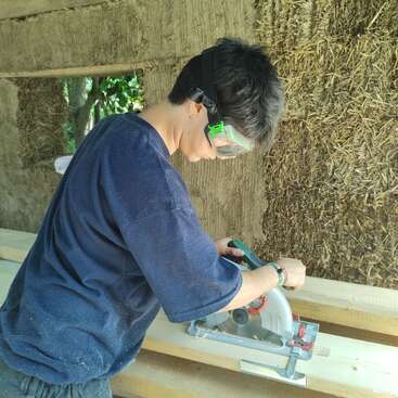 Une personne portant des lunettes de protection utilise une scie circulaire pour couper une planche de bois à l'intérieur d'une structure rustique avec des murs en bottes de paille et des poutres en bois.