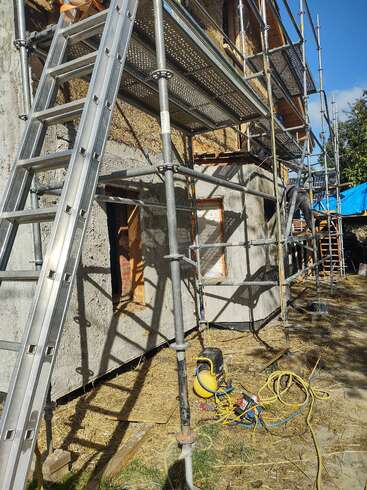 Eine Baustelle mit Metallgerüsten und Leitern umgibt ein Gebäude, das renoviert wird. Man sieht Werkzeuge, Stromkabel, Stroh und unfertige Außenwände im hellen Sonnenlicht.
