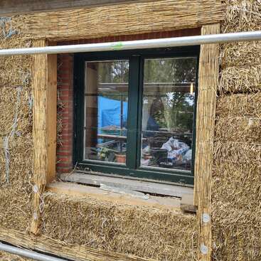 This image shows a window set in a wall made of straw bales and wooden supports, indicating an eco-friendly or natural building under construction or renovation.