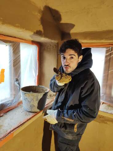 Ein Mann in Arbeitskleidung und Handschuhen hält eine Handvoll Schlamm oder Gips in der Hand. Er steht neben einem Fenster mit Plastikabdeckung und einem schmutzigen Eimer.