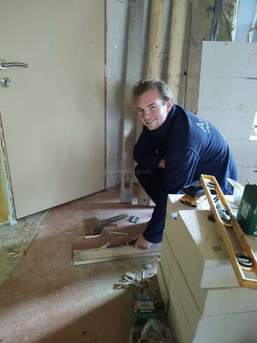 Ein Mann kniet auf dem Boden in einem halbfertigen Raum und arbeitet mit Holz und Werkzeugen. Er lächelt in die Kamera, umgeben von Baumaterialien.