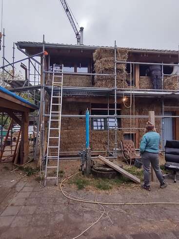 Ein im Bau befindliches zweistöckiges Haus mit Strohballenwänden, Gerüsten, Leitern und Arbeitern. Eine Person in einer blauen Jacke beobachtet draußen den Baufortschritt.