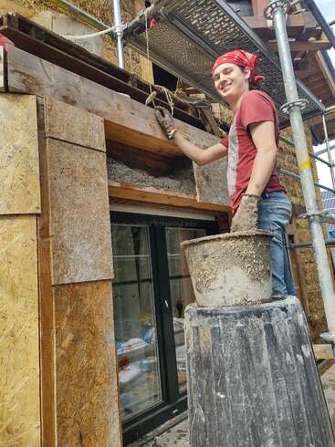 Eine Person mit rotem Halstuch und Handschuhen lächelt, während sie an einem im Bau befindlichen Haus arbeitet und über einem Fenster Putz aufträgt, umgeben von Holzbrettern und Gerüsten.