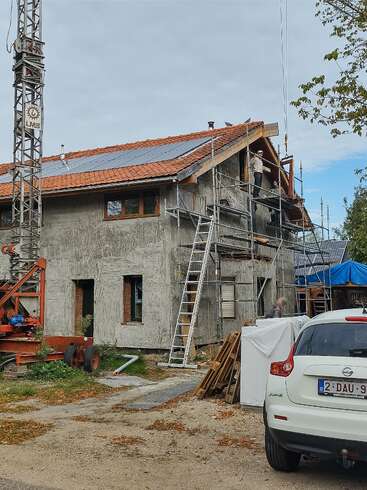 Ein im Bau befindliches Haus mit Gerüsten, Arbeitern, Sonnenkollektoren auf dem Dach, Baumaterialien, einem weißen Auto, einem Kran und Bäumen in der Nähe unter einem bewölkten Himmel.