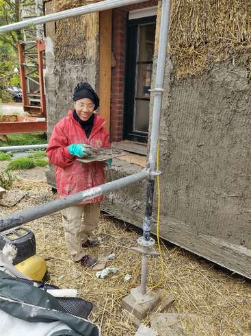 Eine Person, die Handschuhe, eine Schutzbrille und eine rote Jacke trägt, verputzt auf einer Baustelle, umgeben von Gerüsten und Baumaschinen, eine mit Stroh verkleidete Wand.