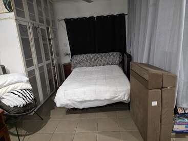 Ein gemütliches Schlafzimmer mit einem Bett in der Mitte, weißer Bettwäsche, großen Schränken auf der linken Seite, Vorhängen dahinter, einem Stuhl mit Leinen und Büchern, die daneben gestapelt sind.