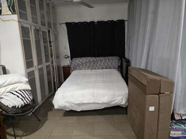Ein gemütliches Schlafzimmer mit einem Doppelbett, einer weißen Bettdecke, einem gemusterten Kissen, schwarzen Vorhängen, einem Kleiderschrank, einem ausklappbaren Sofa, einem Sessel mit Decken und einem Bücherregal. Ruhige Atmosphäre.