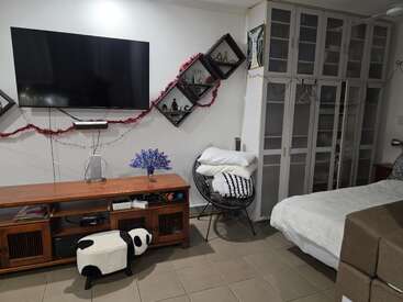 Ein gemütliches Schlafzimmer mit einem an der Wand montierten Fernseher, einem Holzschrank, einzigartigen schrägen Regalen, einem Kleiderschrank mit Glastüren, einem bequemen Sessel mit Kissen und einem verspielten Fußhocker im Panda-Design. Aufgeräumt und einladend.