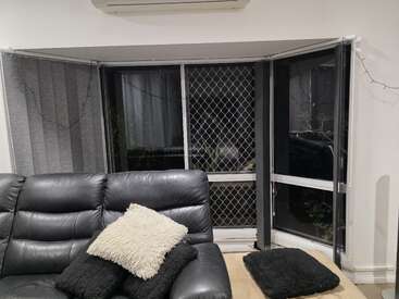 Eine gemütliche Wohnzimmerecke mit einem schwarzen Ledersofa, flauschigen schwarz-weißen Kissen, großen Erkerfenstern mit Fliegengittern, Vertikaljalousien und Lichterketten.