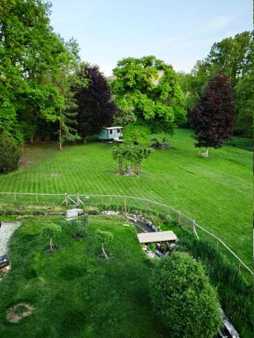 Un jardin verdoyant à la pelouse impeccablement entretenue, entouré d'arbres. Un petit pont en bois enjambe un ruisseau, et une petite maison se niche sous des arbres feuillus.