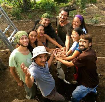 The image depicts a group of nine people, including a dog, standing in a circle with their hands clasped together, posing for a photo in a dirt area.